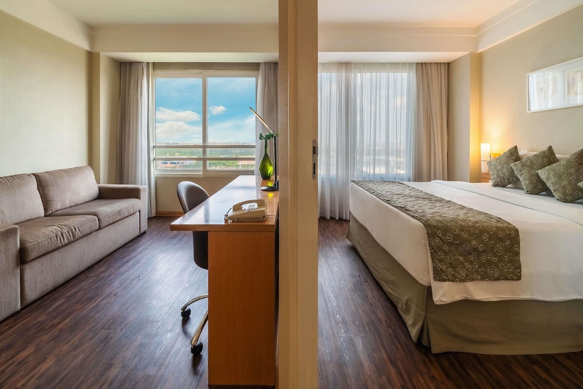 Habitación de hotel con cama, sofá, escritorio con teléfono y ventana con cortinas.