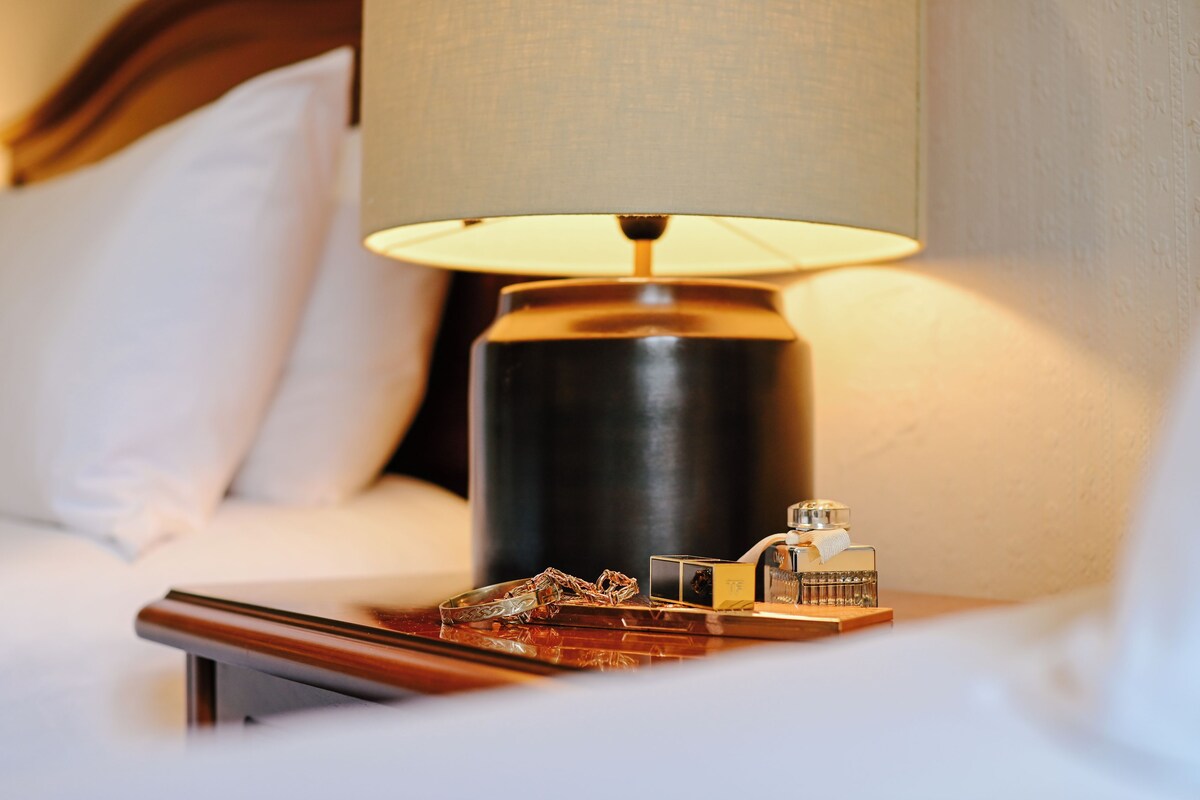 Habitación de hotel con mesita de noche, lámpara, un reloj pequeño y una bandeja con aperitivos.