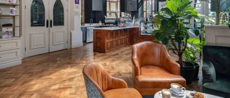 Una habitación amplia con suelo de madera en patrón espiga, una planta grande en maceta, sillones de cuero y una mesa con una taza de café y pasteles.