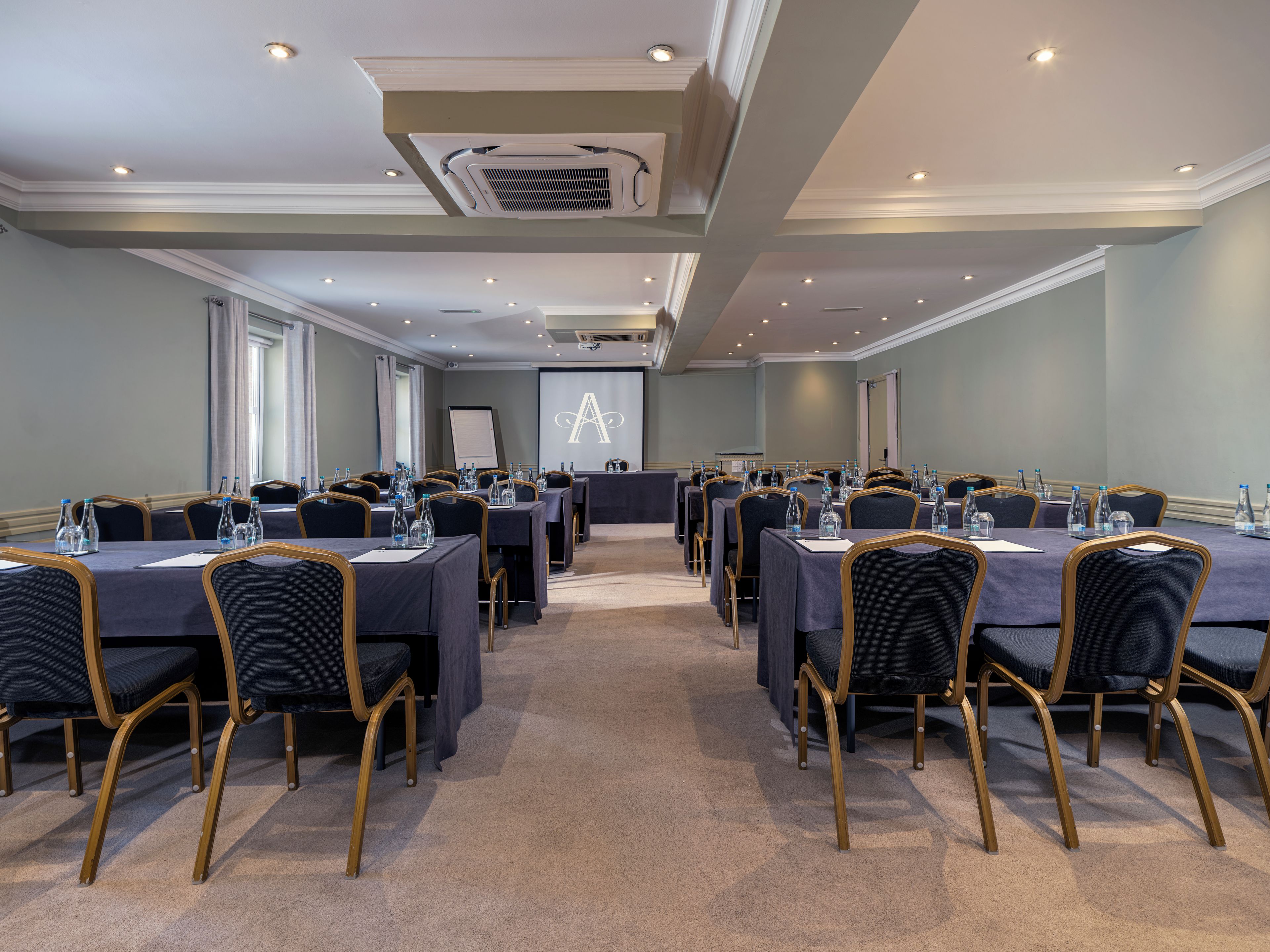 A conference room with multiple tables and chairs, equipped with water bottles and a projector screen.