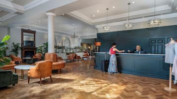 A hotel lobby with a reception desk, leather seating, a herringbone wood floor, and a large mirror above the fireplace.