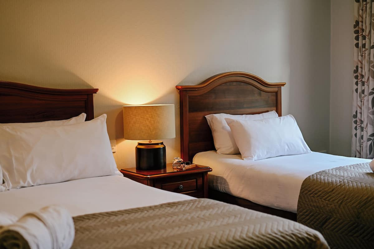A hotel room with two beds, a nightstand with a lamp, and a window with curtains.