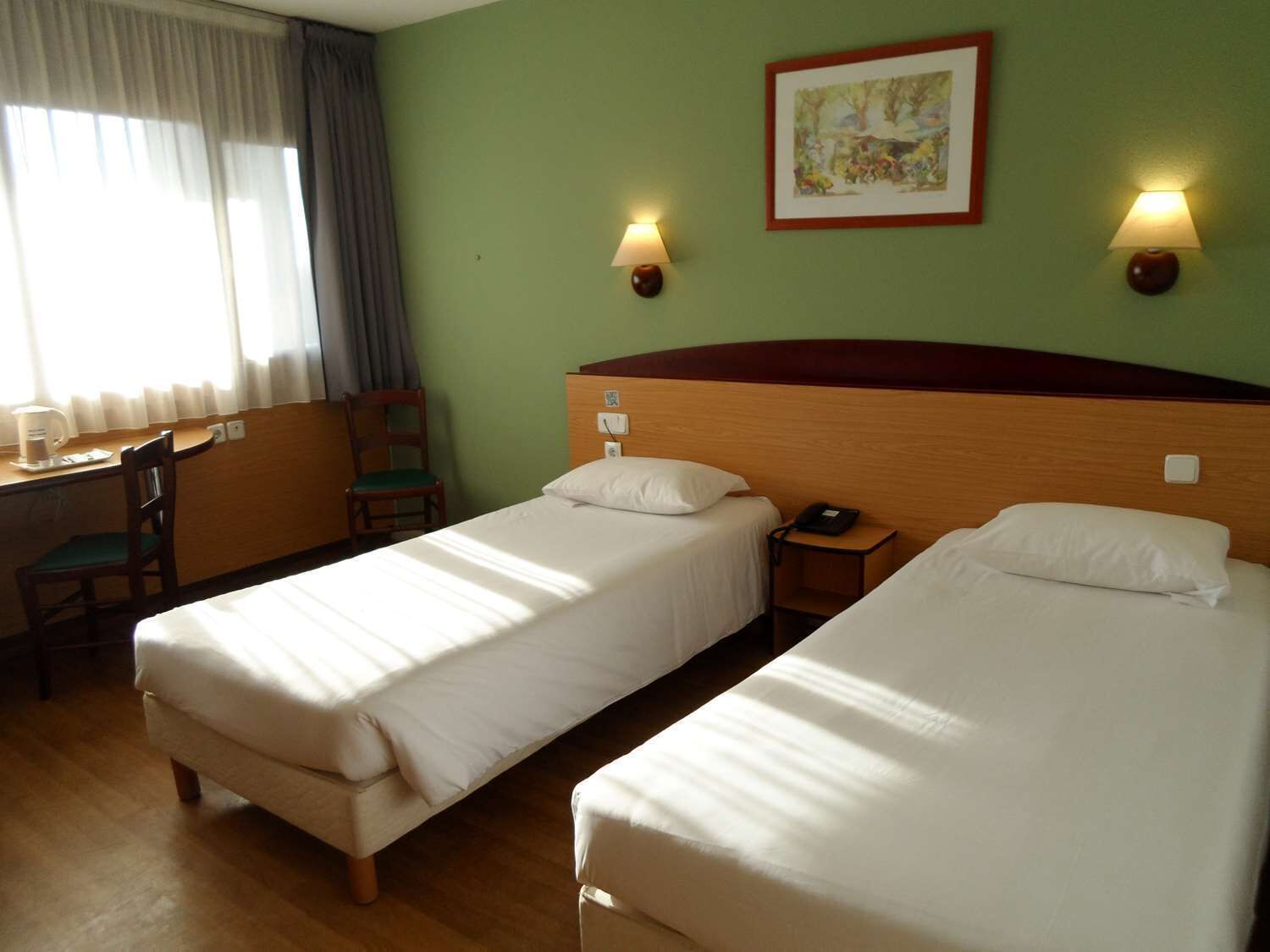 Une chambre d’hôtel avec deux lits simples, un bureau avec une chaise, une machine à café et un tableau au mur.