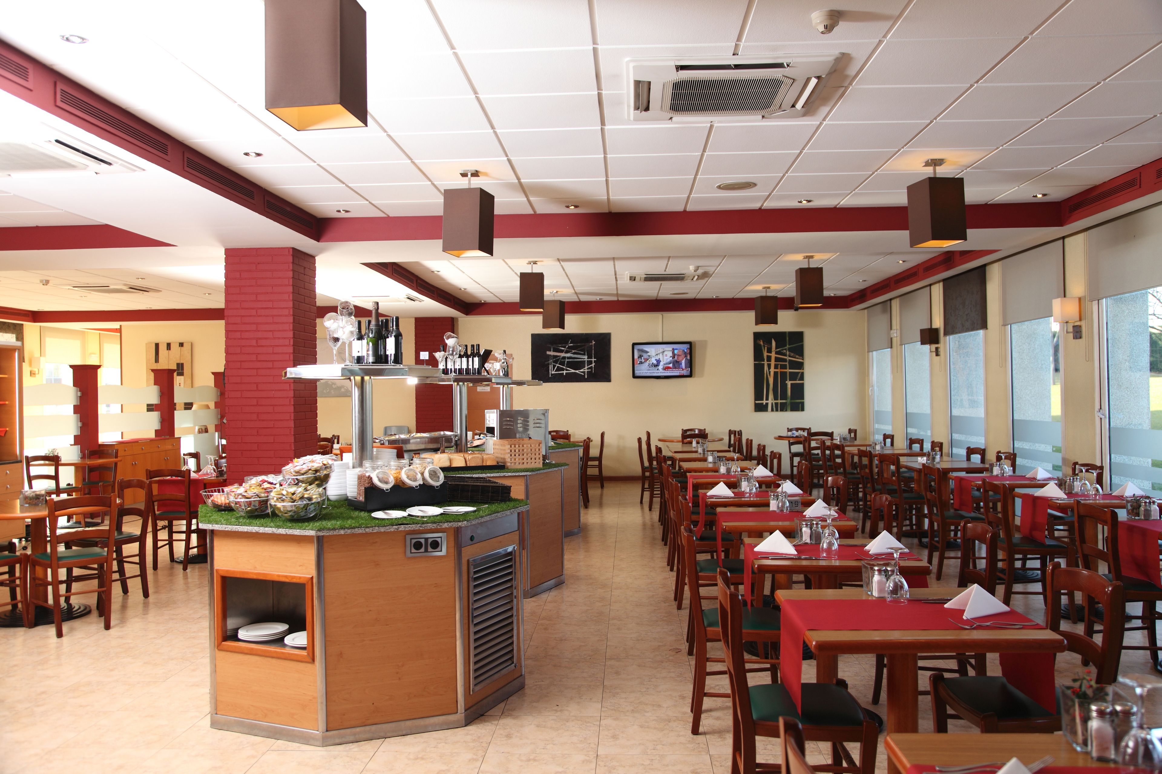 A restaurant with multiple dining tables and chairs, a central counter with food items, and a television mounted on the wall.