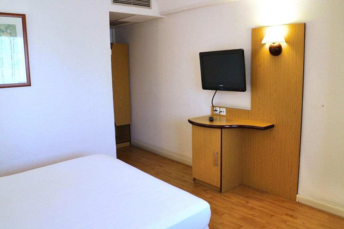 A hotel room with a bed, a wooden desk, a flat-screen TV mounted on the wall, and a bedside lamp.