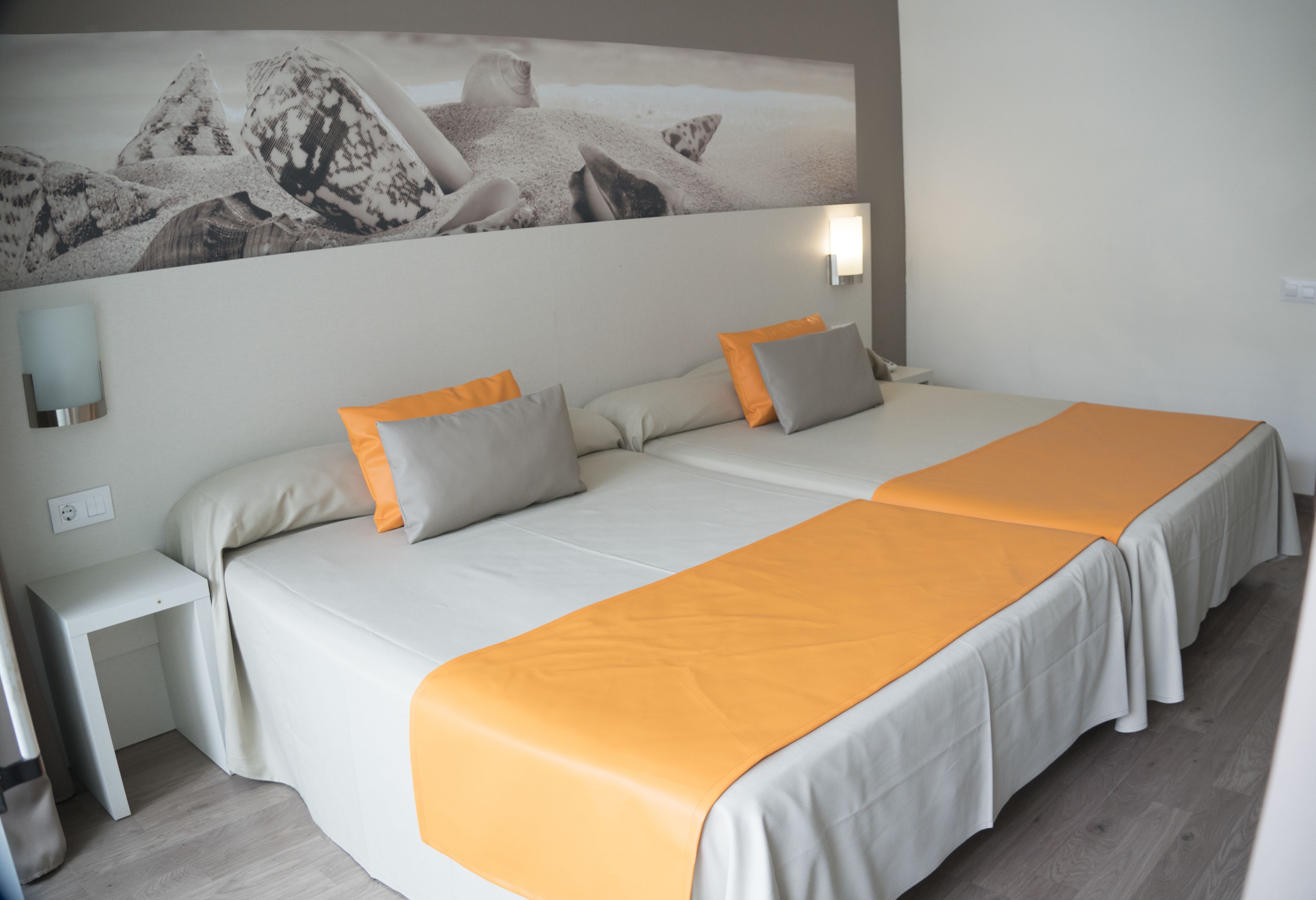 A double bed with an orange bedspread, two pillows, a nightstand, a wall-mounted light, and a large abstract painting above the bed.
