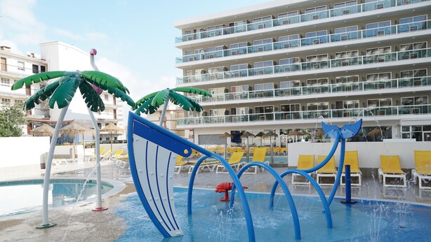 Ett poolområde med vatteninstallationer, solstolar och en byggnad med balkonger i bakgrunden.