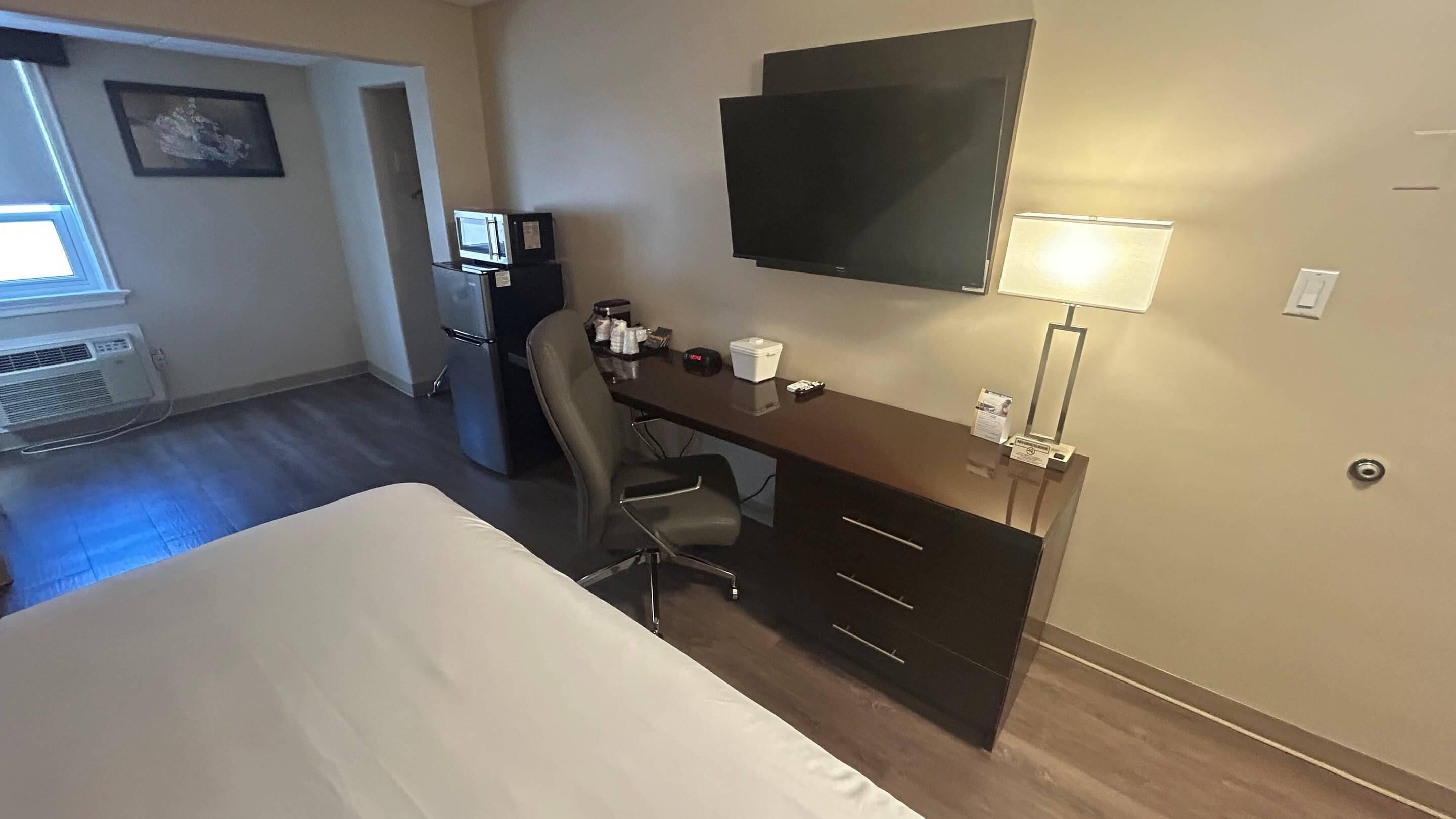 A hotel room with a bed, desk, chair, TV, and a small kitchenette.