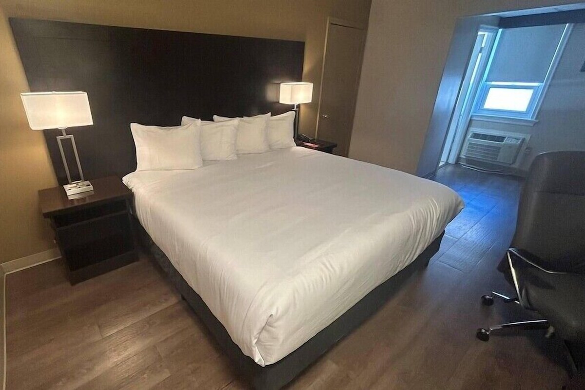 A hotel room with a large bed, two bedside tables with lamps, a chair, and a window with blinds.