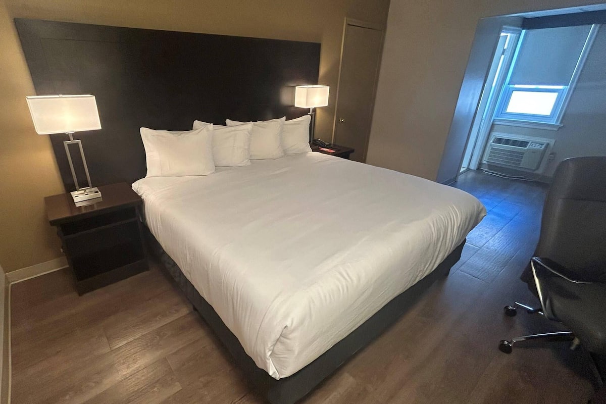 A hotel room with a large bed, two bedside tables with lamps, a chair, and a window with blinds.