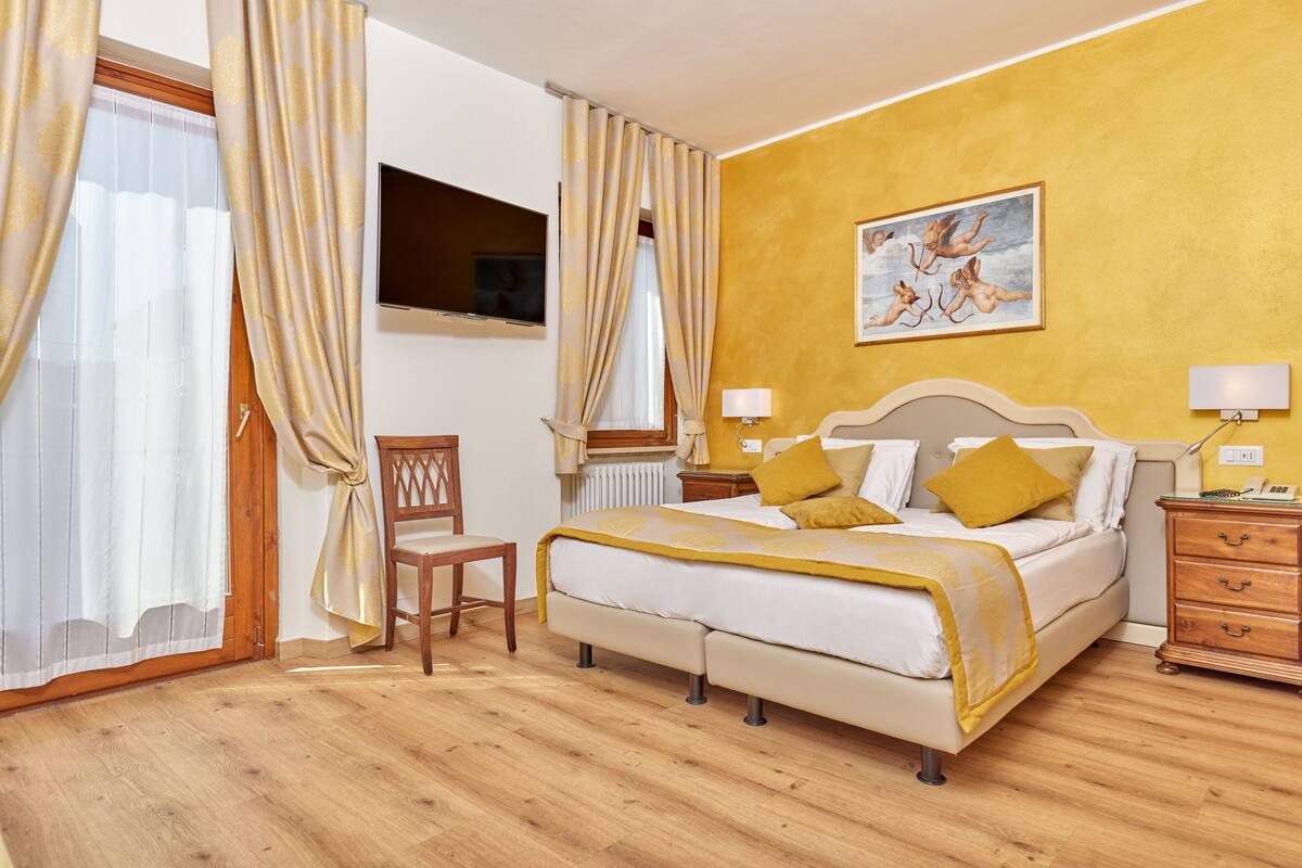 Una habitación de hotel con una cama, mesitas de noche, una silla, un cuadro en la pared y cortinas.