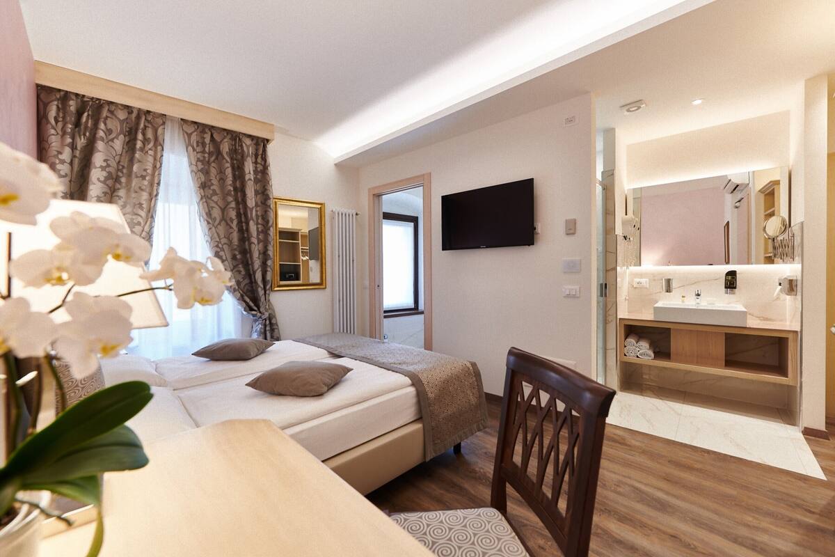 Habitación de hotel con cama, escritorio, silla, televisor, baño con lavabo y espejo, y ventana con cortinas.