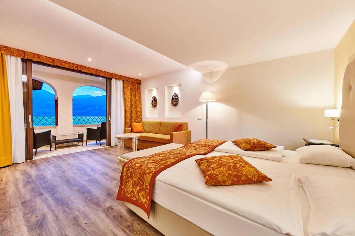 Habitación de hotel con una cama grande, un sofá, una mesita y vistas a las montañas a través de una puerta abierta.
