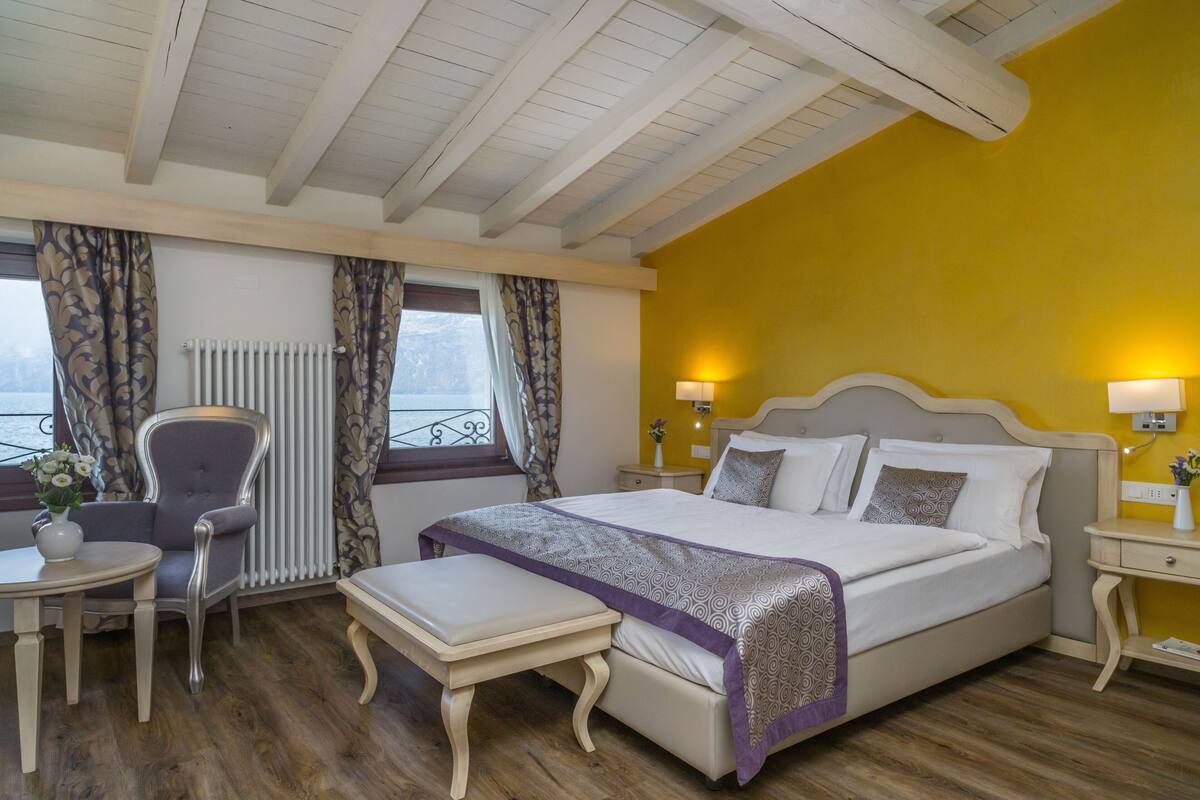 Un dormitorio con cama, mesitas de noche, una silla, un banco, una pequeña mesa con un florero y una ventana con cortinas.