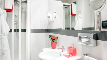 Ein modernes Badezimmer mit Duschkabine aus Glas, einem weißen Waschbecken mit einer rosa Blumenvase und einem Spiegel mit rotem Handtuch.