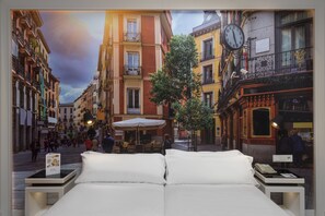 A bed with white linens in a hotel room with a view of a street with buildings and a clock. - Elba Madrid Alcalá (Madrid)