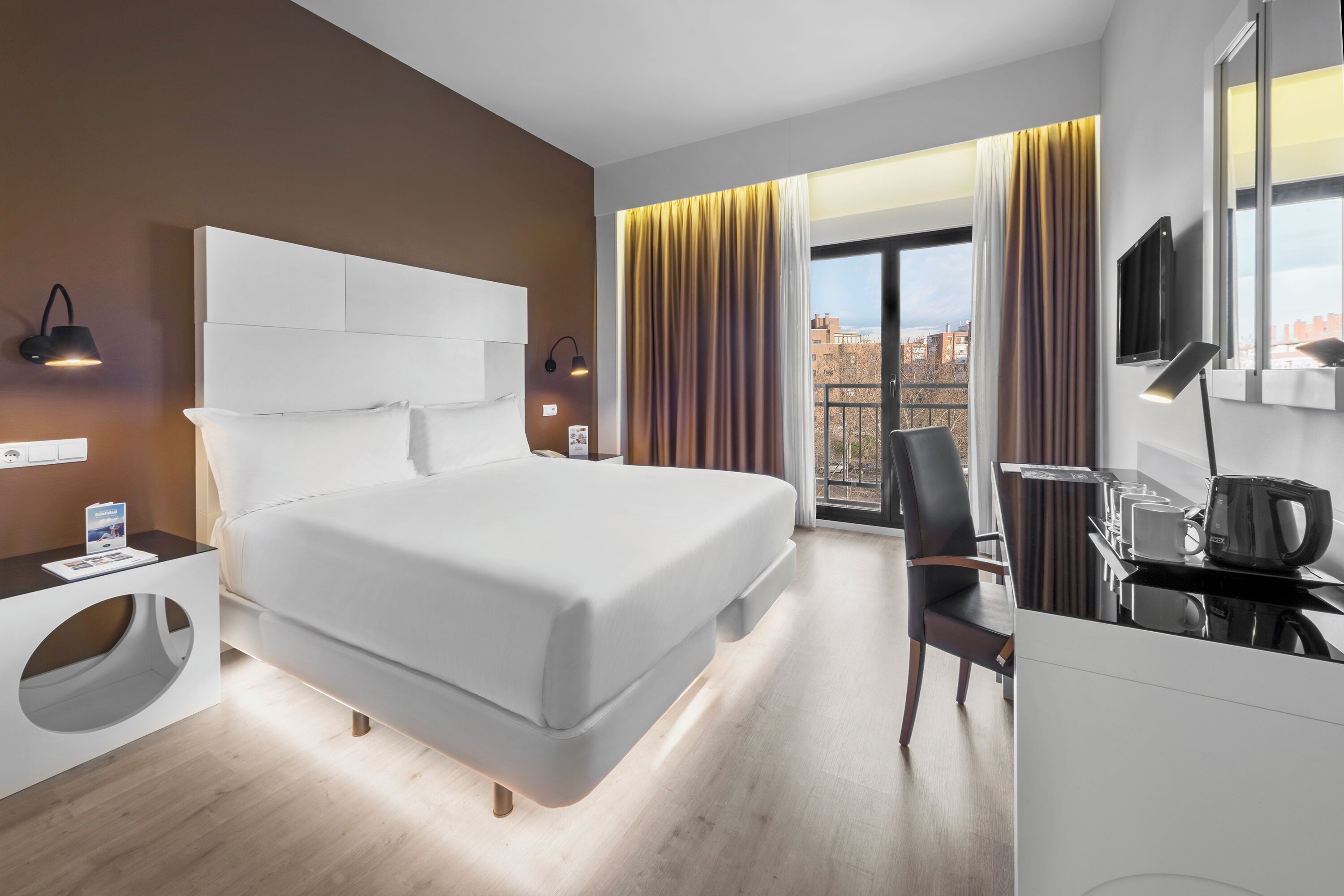 Una camera d'albergo moderna con un letto grande, una scrivania con bollitore, una sedia e vista sulla città da ampie finestre.