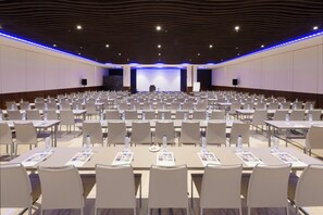 A conference room with rows of tables and chairs, a stage with a screen, and a modern ceiling design. - Elba Madrid Alcalá (Madrid)