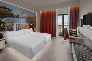 A hotel room with a bed, a desk, a chair, a TV, and a view of a building through the window. - Elba Madrid Alcalá (Madrid)