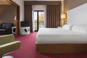 A hotel room with a bed, a television, a seating area with a chair and a sofa, a small table, and a balcony with curtains. - Elba Madrid Alcalá (Madrid)
