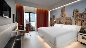 Una moderna camera d'hotel con un letto grande, una scrivania con una sedia, una TV a schermo piatto e un murale raffigurante un edificio storico.