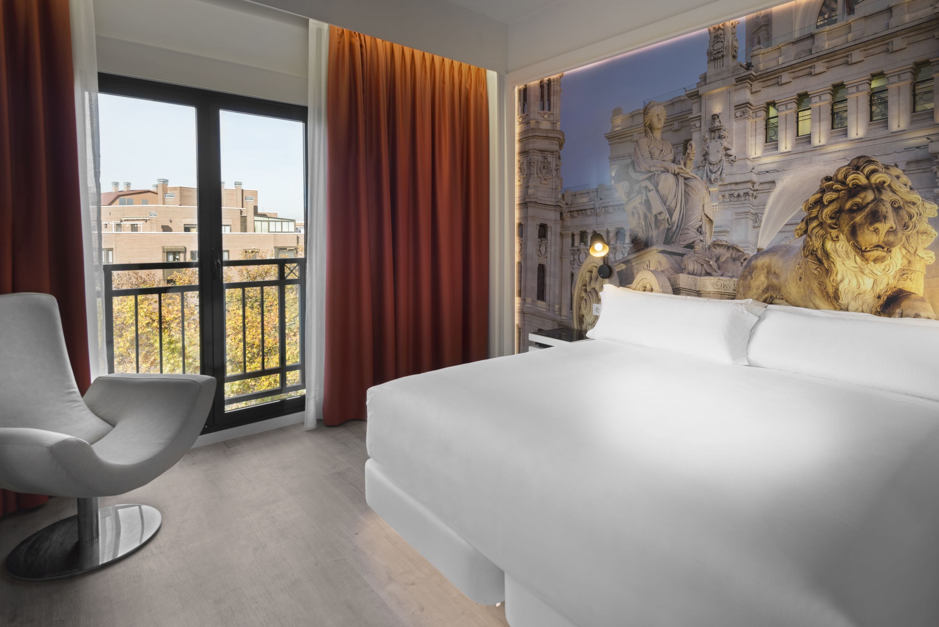 A hotel room with a large bed, a modern chair, a view of buildings, and a mural of a lion.