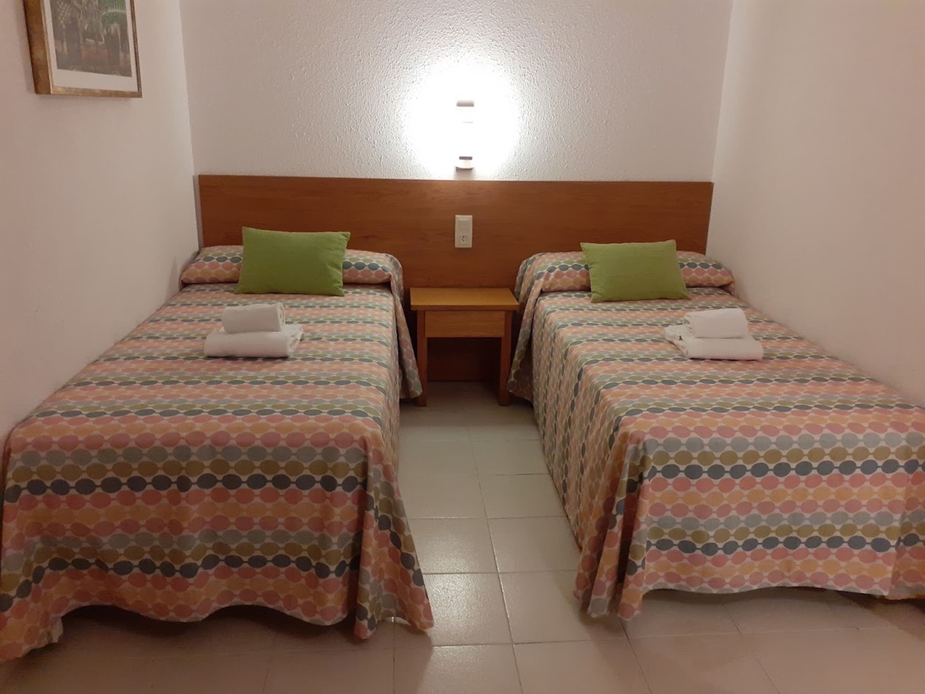 Una habitación con dos camas individuales, cada una con una colcha estampada y un almohátero verde. Hay un cabecero de madera entre las camas y una mesita de noche de madera con una lámpara.
