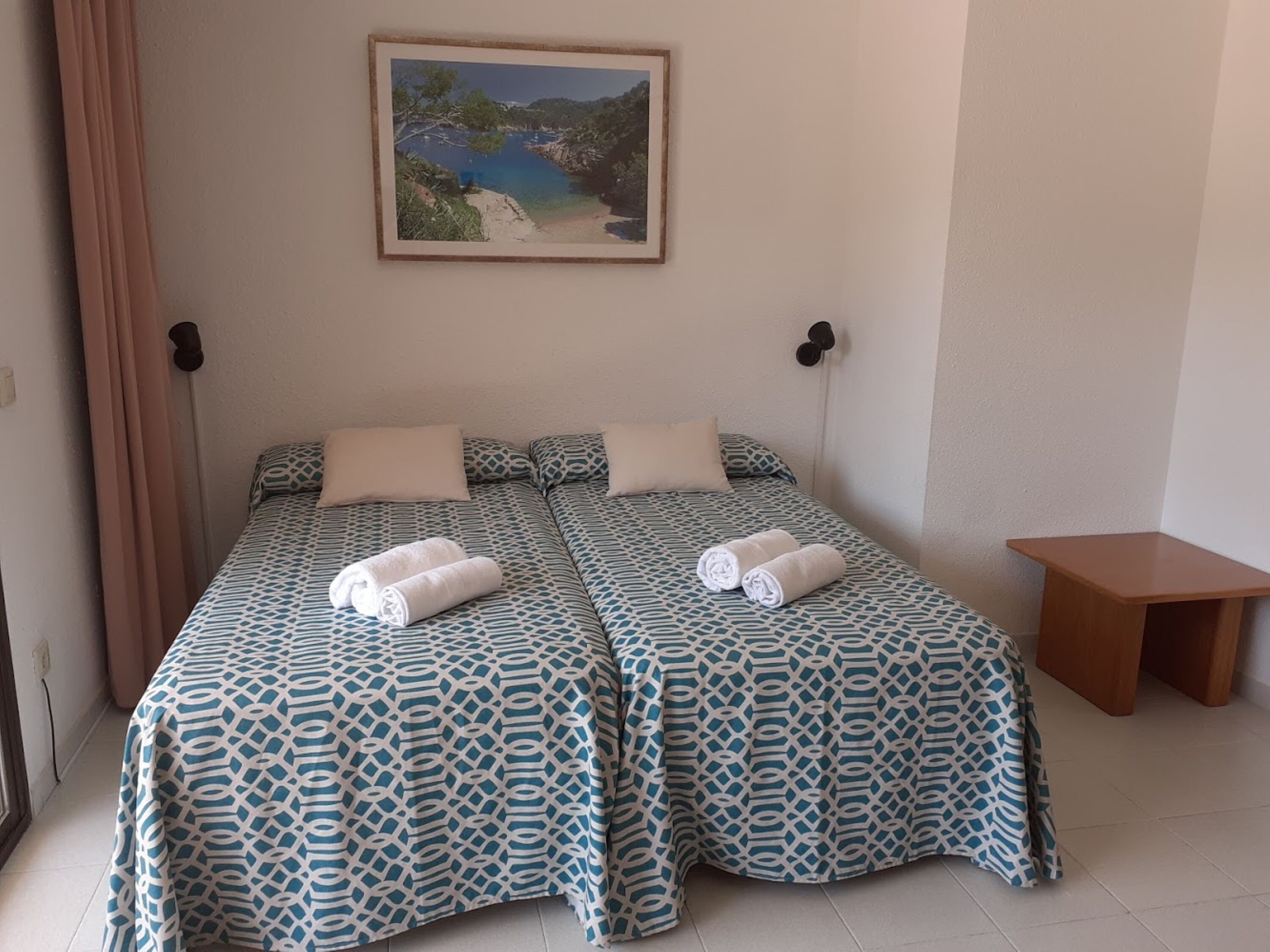 Un lit double avec une literie à motifs, une petite table de chevet et un tableau représentant un paysage côtier accroché au mur.