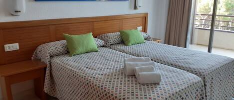 Habitación doble con dos camas, cada una con ropa de cama estampada y almohadas verdes. Hay una mesita de noche con una lámpara, un cuadro en la pared y vista al exterior a través de una ventana.