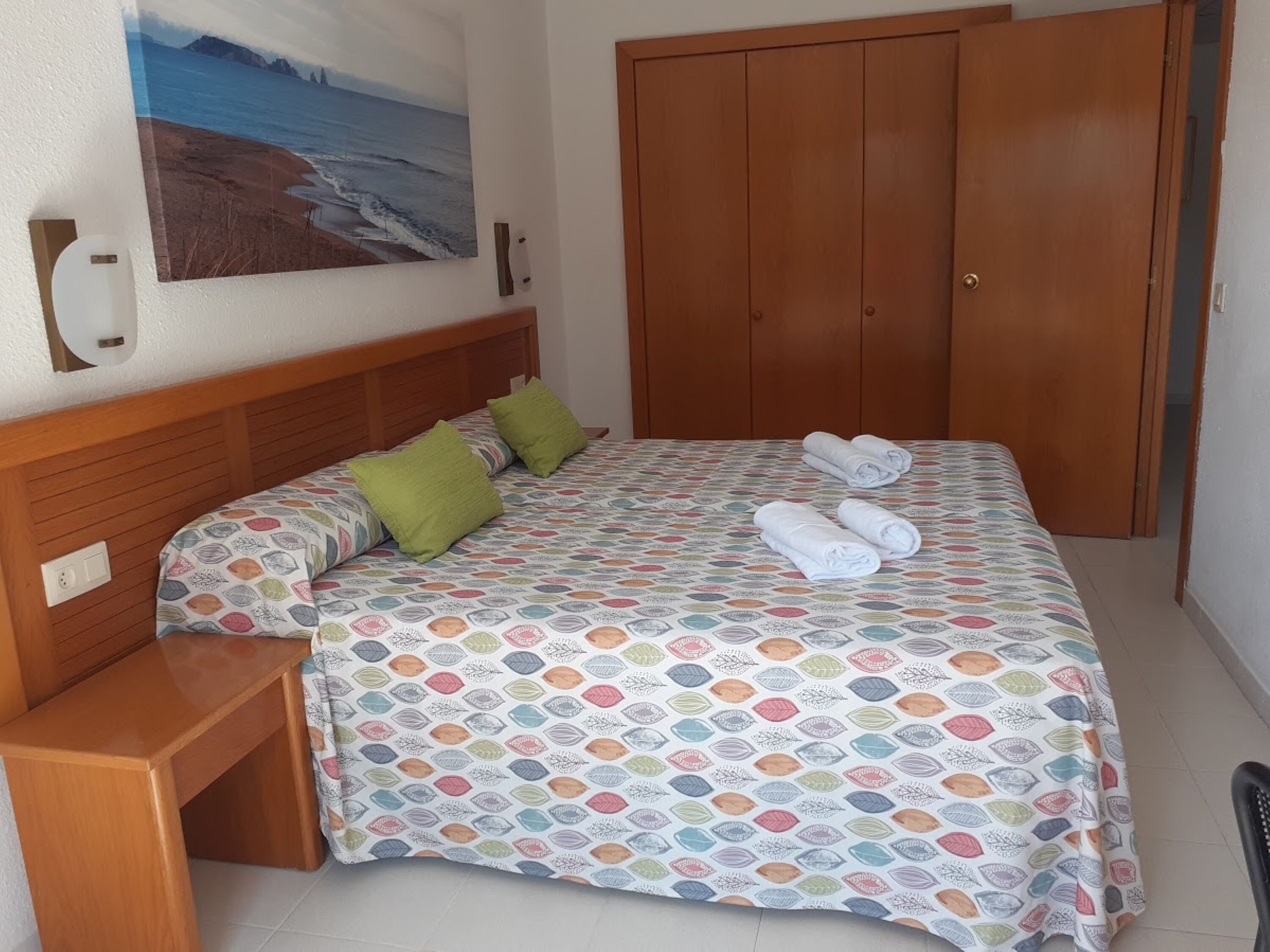 A bedroom with a bed, two pillows, a wooden nightstand, a wooden wardrobe, and a painting of a beach scene on the wall.