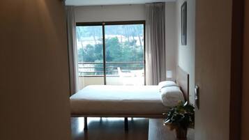 Camera d'albergo con un letto, un comodino con una pianta, una finestra che offre vista su alberi e edifici, e una lampada a parete.