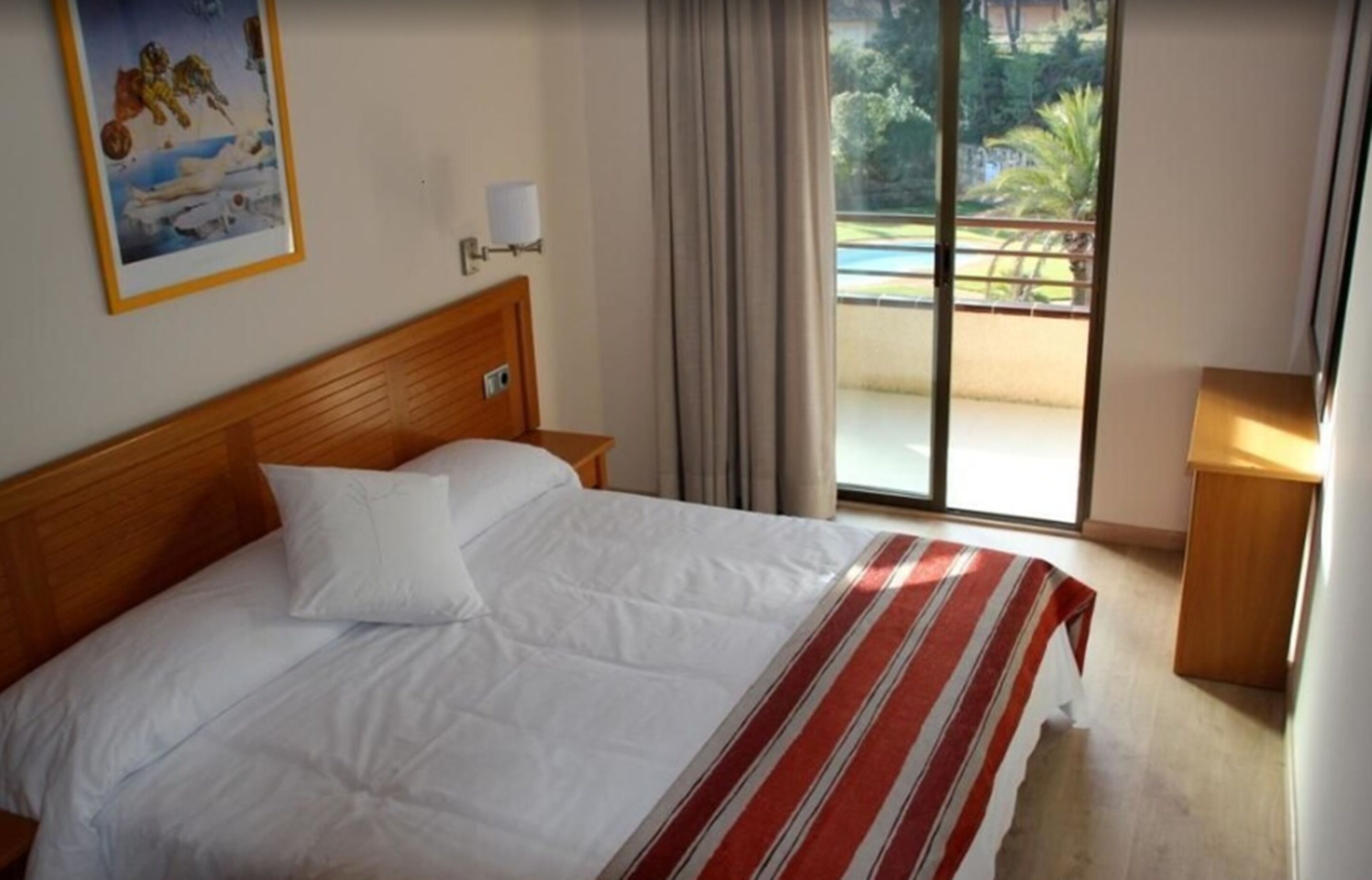 Habitación de hotel con una cama, una mesita de noche, una lámpara, un cuadro y vista a una piscina por una puerta corredera.