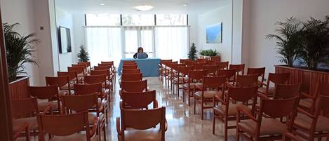 A conference room with rows of wooden chairs facing a blue table where a person is seated.