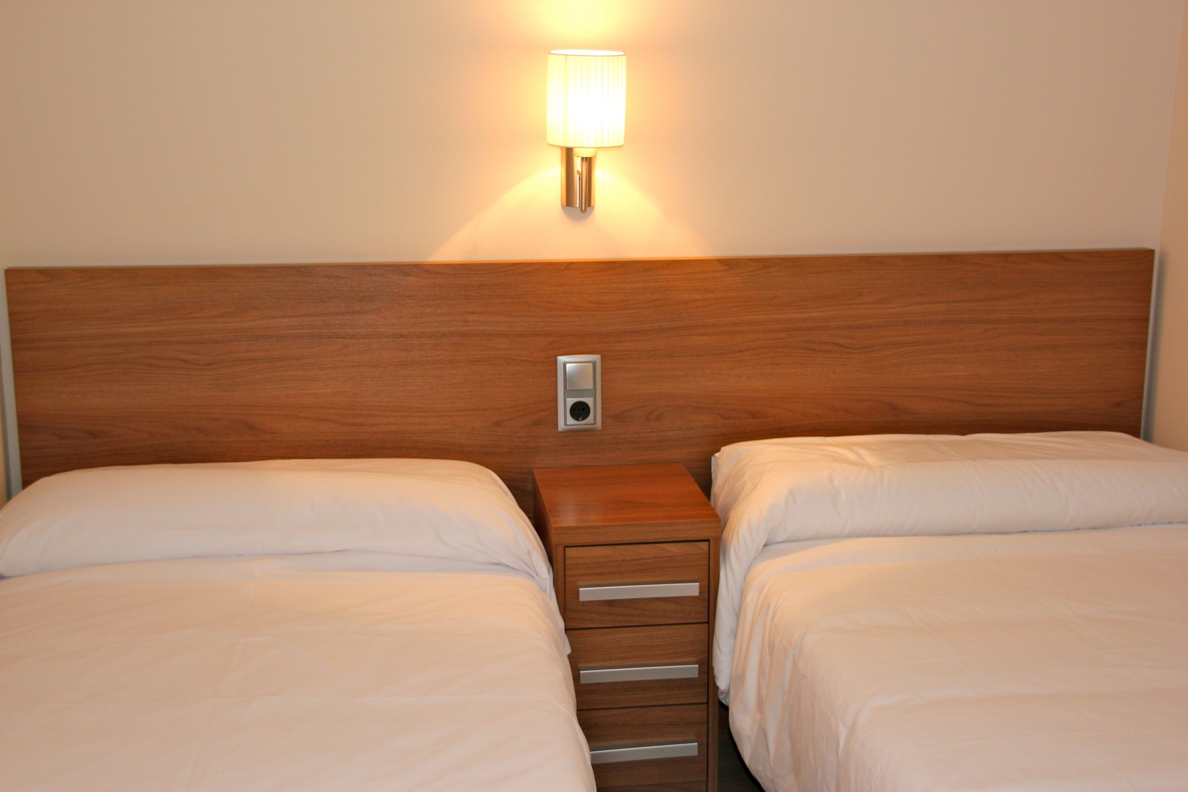 A hotel room with two beds, a wooden headboard, a bedside table with drawers, and a wall-mounted lamp.