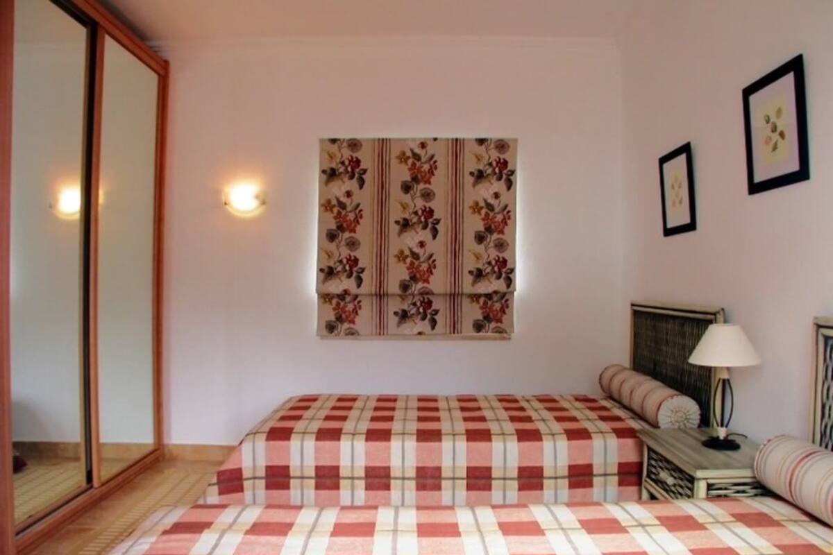 A bedroom with a checkered bedspread, a wall-mounted floral tapestry, a bedside lamp, and a mirror.
