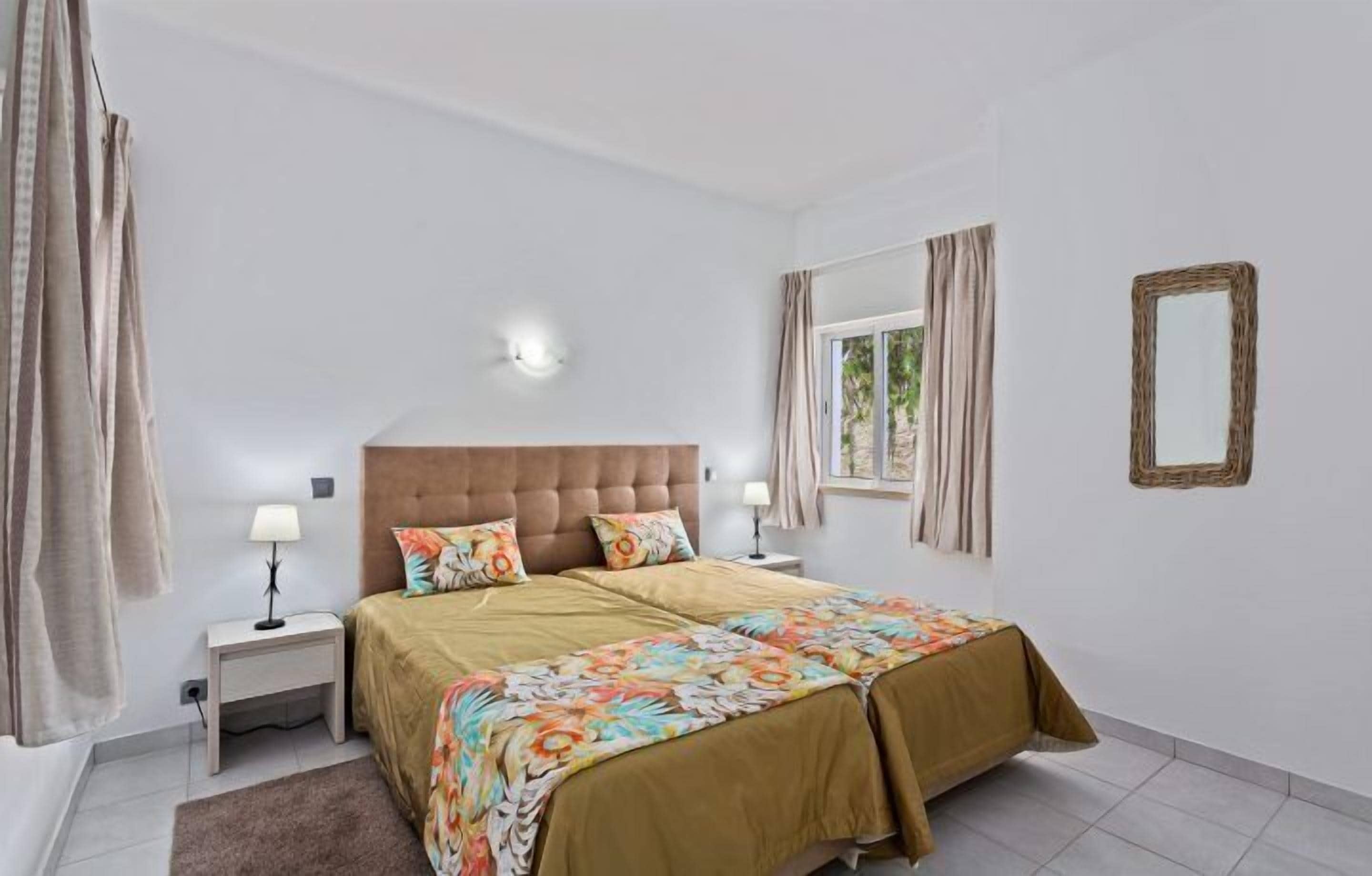A bedroom with a large bed, two bedside tables, a window with curtains, and a wall-mounted mirror.