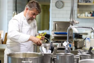 Un chef, vêtu d’un uniforme blanc, prépare des plats dans une cuisine équipée de casseroles en acier inoxydable et d’une horloge murale.