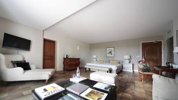 Una sala de estar con un sofá, un sillón, una mesita de centro con revistas, un televisor y una cama al fondo.