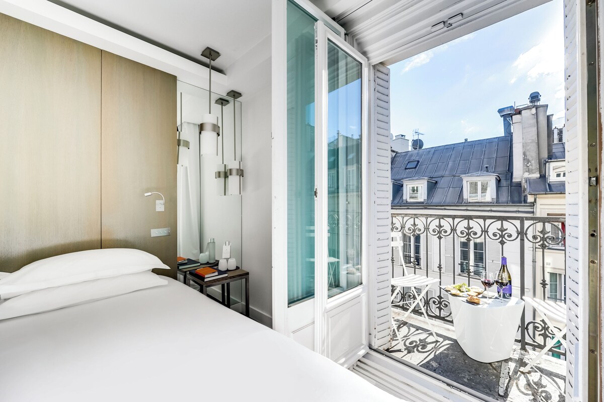 Een moderne hotelkamer met een groot bed, een nachtkastje met een lamp, en een balkon met een eettafel.