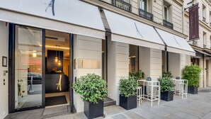 A hotel entrance with a glass door, outdoor seating area with tables and chairs, and potted plants. - Hotel Le A (Paris)