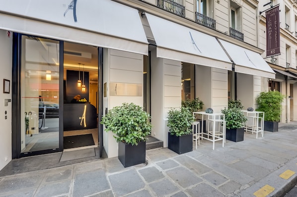 Entrada de hotel com porta de vidro, área de estar externa com mesas e cadeiras e plantas em vasos.
