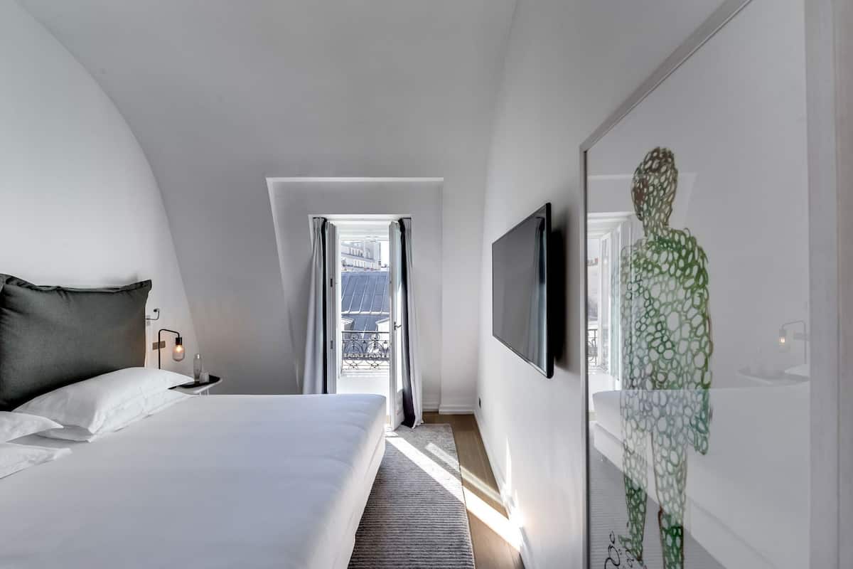 A modern hotel room with a bed, a TV, and a view of the city through a window.