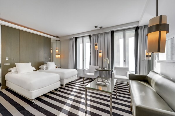 Una habitación de hotel moderna con dos camas, un sofá, una mesa de centro de vidrio y ventanas grandes con cortinas.