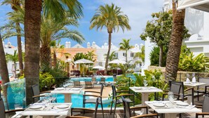 Un área de comedor al aire libre con mesas y sillas, una piscina y palmeras.