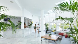 Un hall de hotel espacioso y moderno con suelo de mármol, un mostrador de recepción, una zona de estar con sofá y mesa de centro, y ventanas amplias con vistas al exterior.