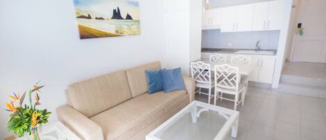 A modern living room with a beige sofa, a glass coffee table, and a dining area with white chairs and a white cabinet.