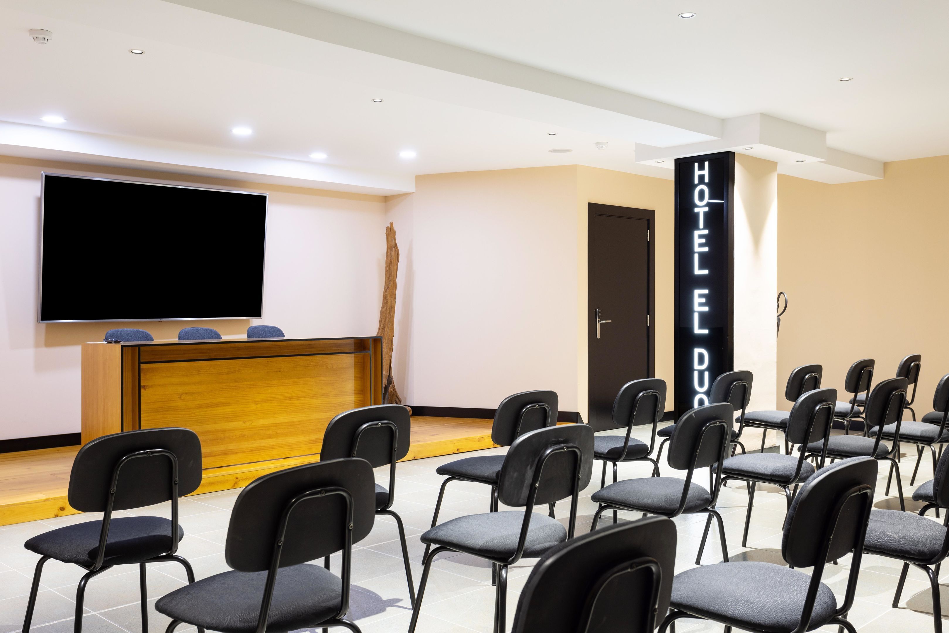 A hotel conference room with rows of black chairs, a wooden reception desk, a large flat-screen TV, and a 'HOTEL DUO' sign.
