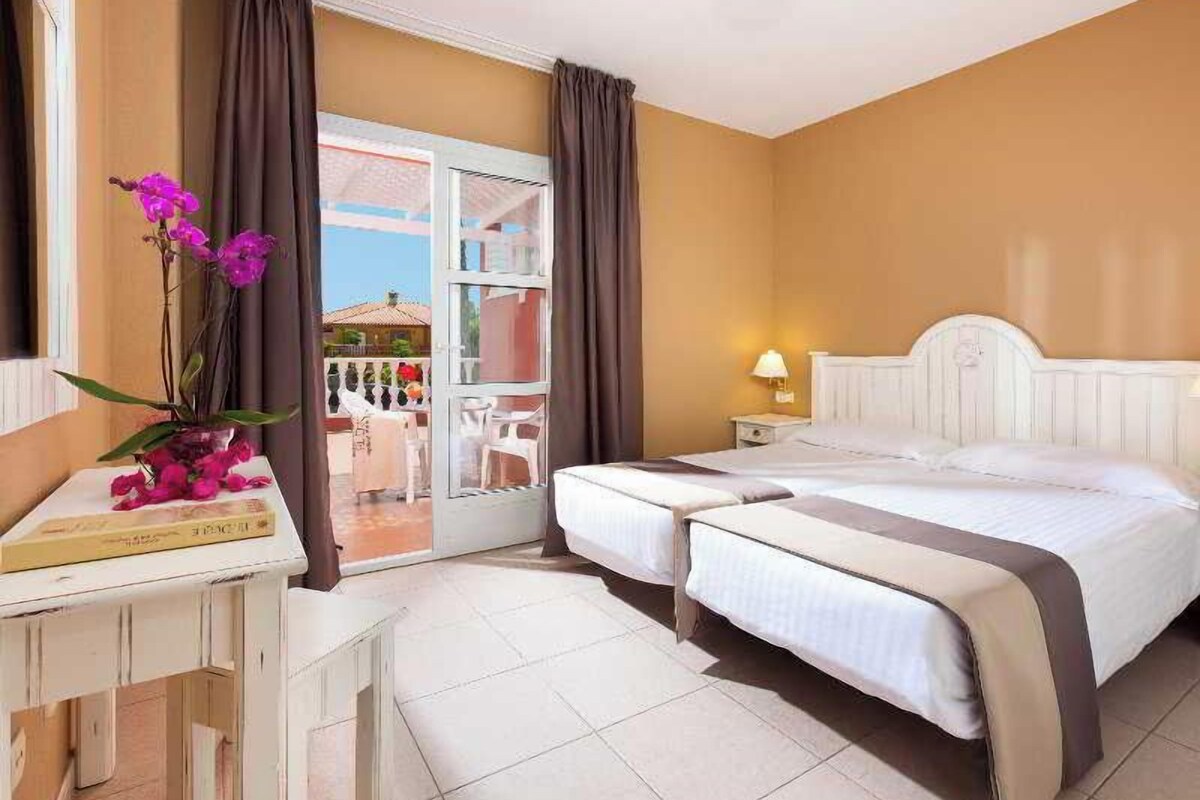 A hotel room with a bed, bedside table, lamp, and a view of the outside balcony.