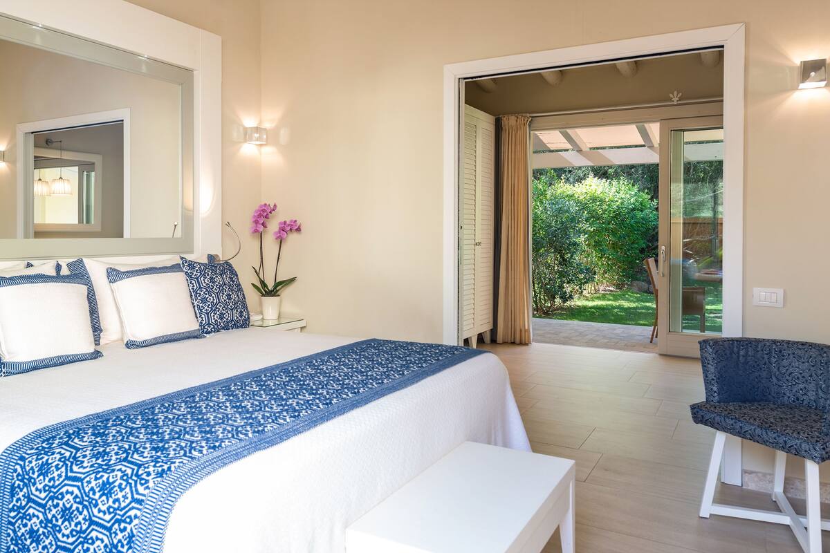 Une chambre à coucher avec un grand lit, une chaise et une vue sur un jardin, visible à travers une porte ouverte.