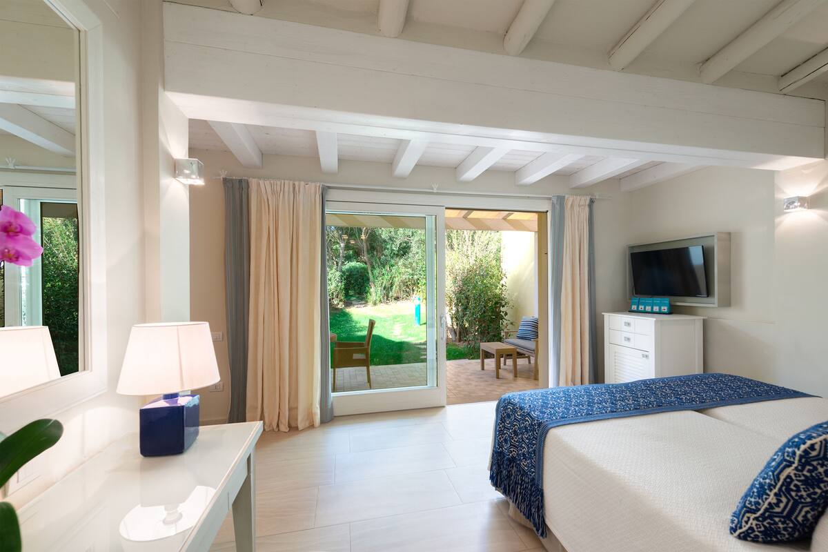 Une chambre d’hôtel avec un lit, une télévision, un bureau avec une lampe et une vue sur le jardin, visible à travers la porte-fenêtre coulissante.